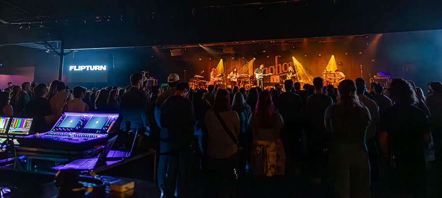 A crowd watches a live performance at the Longhorn Ballroom while sound and lighting equipment operators work from a control area in the foreground. The stage is illuminated with warm concert lighting as musicians perform before an audience filling the venue.