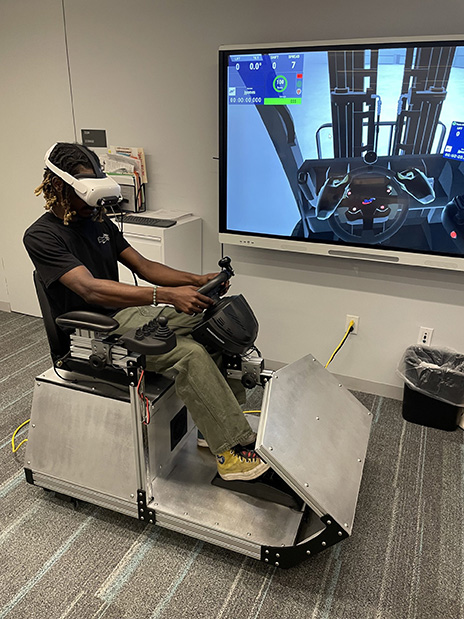 A man is using a forklift simulator that is metal, and has a car steering wheel like set up.