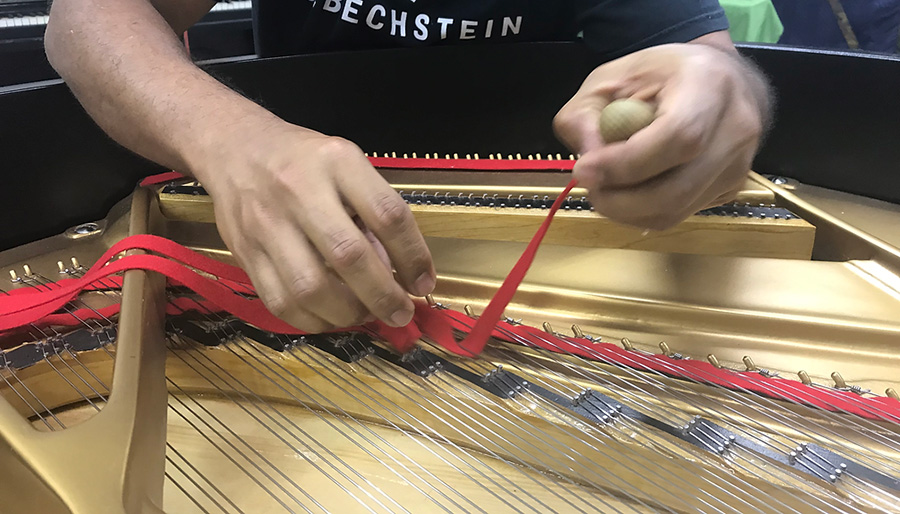 Soto working directly over the piano's interior, threading or installing a bright red felt strip between rows of steel piano strings. Both hands are visible, with one hand guiding the felt and the other holding a wooden-handled tool. The piano's gold-colored cast-iron plate, strings, tuning pins, and wooden action components are visible inside.
