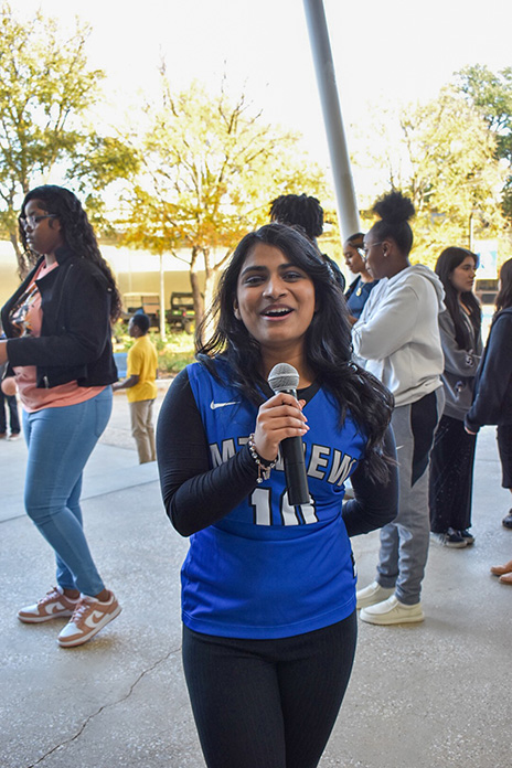 Tiffiya on a Dallas College campus, wearing a blue Mountain View jersey and talking on a microphone enthusiastically. There is a group of Dallas College students in the background.
