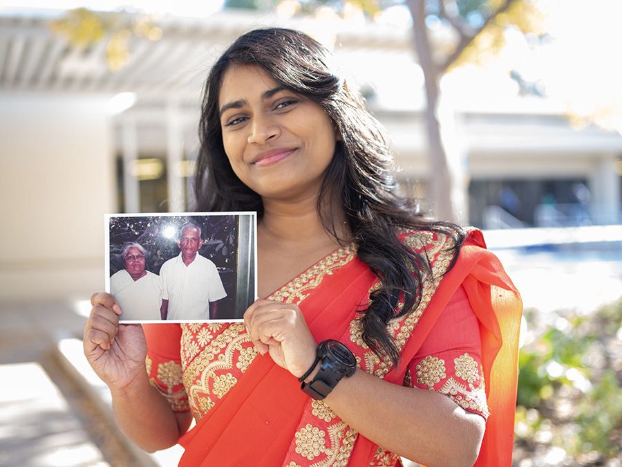 Tiffiya standing on a Dallas College campus, wearing a red sari with a gold pattern. A dark wristwatch is visible on one wrist. She smiles at the camera while holding a photograph with both hands of her grandparents.