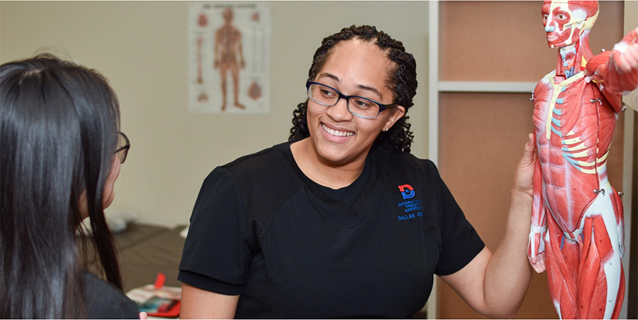 Tometra Williams wearing a black shirt with the Dallas College logo on her upper left corner of the shirt is in a hospital holding a patient bed.