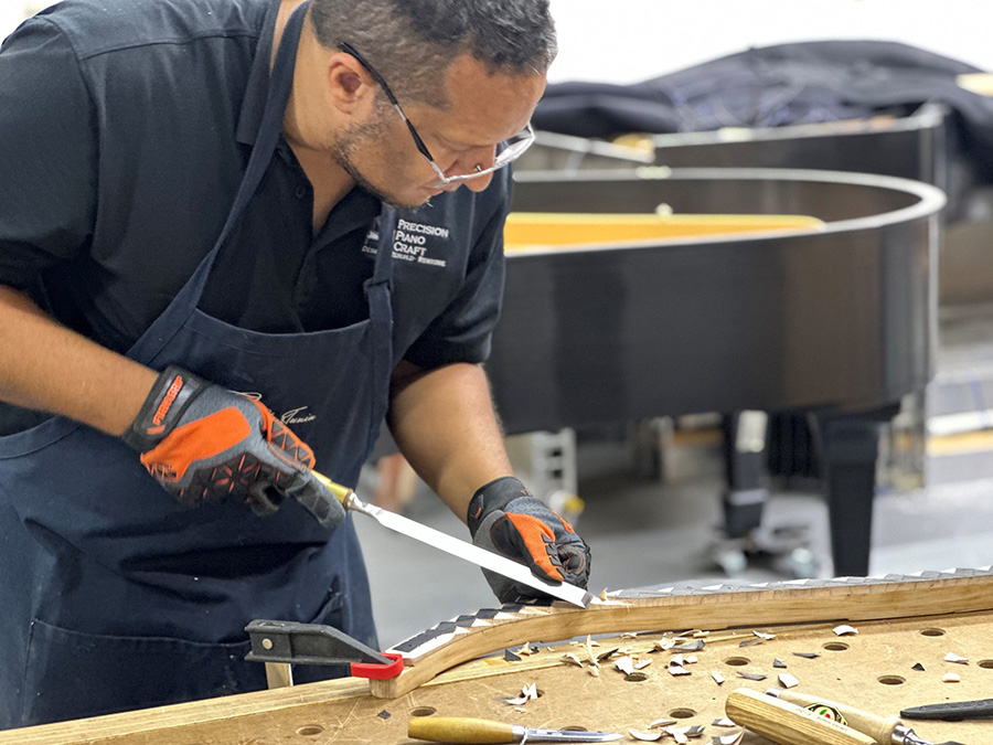Victor Soto working on piano restoration in a workshop. Wearing an apron, safety glasses, and work gloves, Soto uses a hand tool to trim a wooden piano component secured to a workbench, with wood shavings scattered across the surface.
