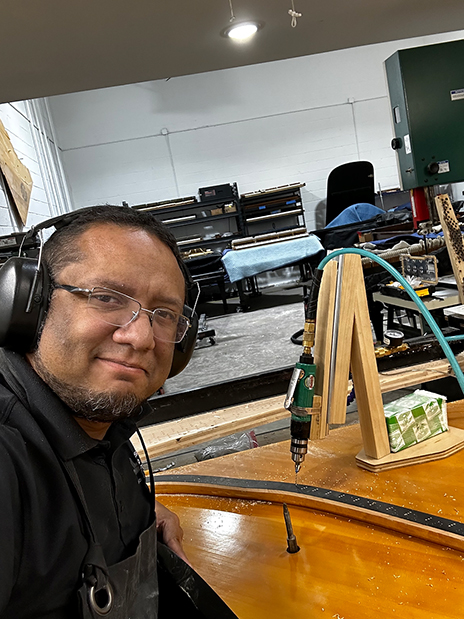 Soto wearing a black apron and black headphones, showing the interior of the piano and the tool he is using. He is also showing his workshop in the background.