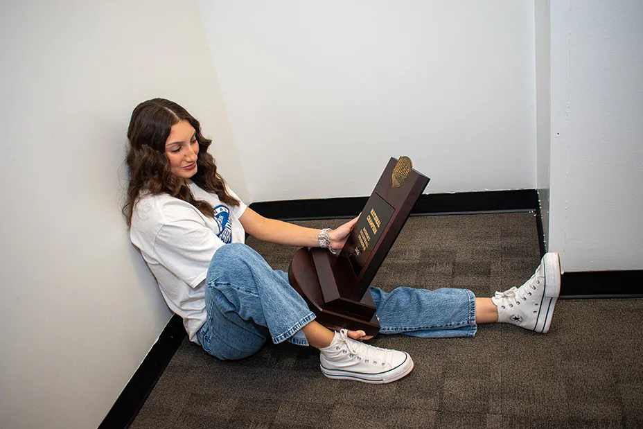 Mia Santos sitting on floor admiring the NJCAA Division III volleyball trophy.
