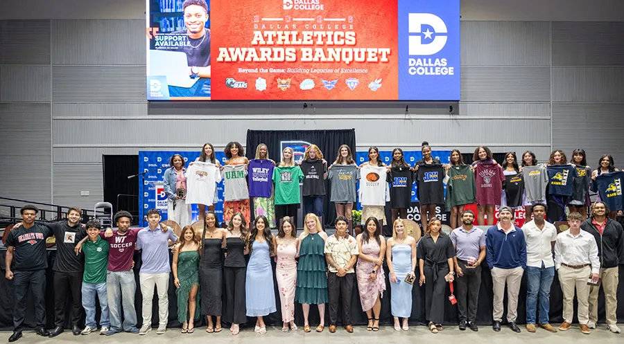 Two rows of student athletes on stage. Top row are holding up college t-shirts.