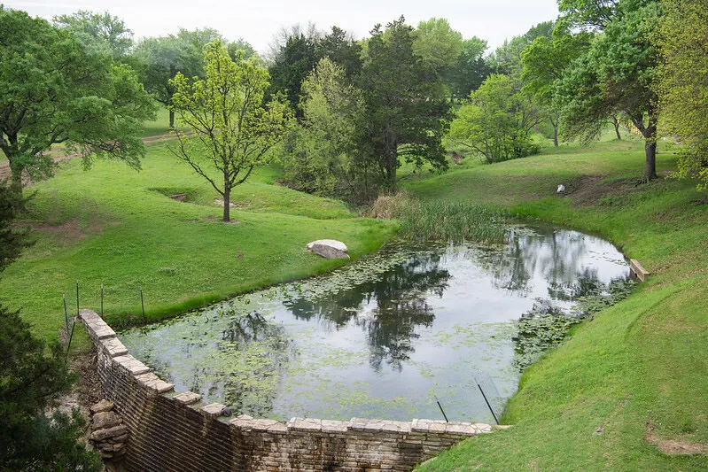 Green area with trees, a pond and a dam.