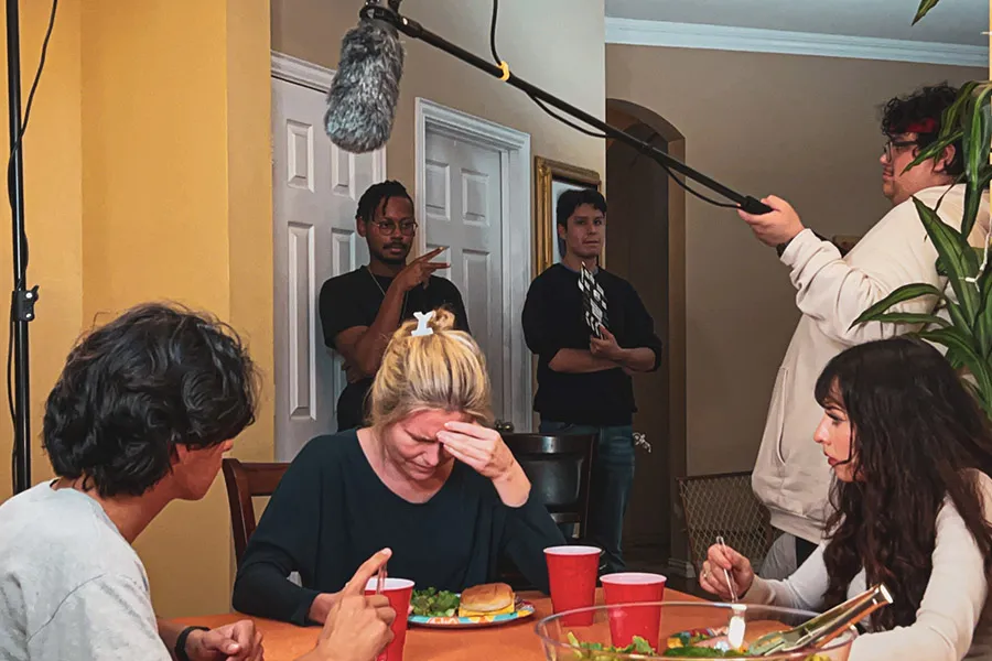 Actors sit at table with filming crew behind them.
