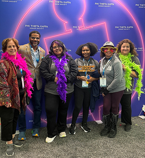 Two administrators and student posing in front of a PTK photo wall.