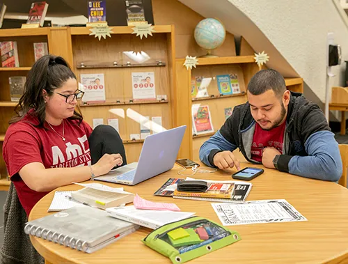 Two students working at a round table in a study room, surrounded by a laptop, notebooks, papers, and stationery.