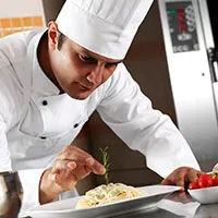 A chef in a white uniform and hat carefully garnishes pasta with rosemary in a kitchen
