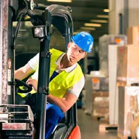 A worker wearing a blue hard hat and yellow vest operates a forklift in a warehouse.