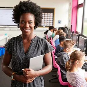A teacher poses for the camera with a notebook in their hand, their class is seen in the background