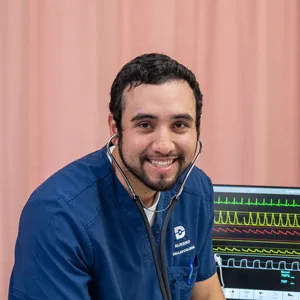 A nursing student sits next to a bed with a monitor running in the background